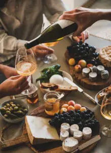 Human hand pouring white orange or rose wine from bottle to glass during party over table with various gourmet snacks cheese grape bread. Gathering, celebrating, wine tasting and party concept